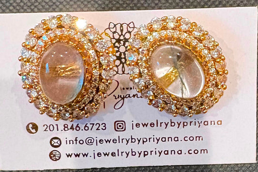 A pair of gold-toned oval stud earrings with clear crystal halo detailing and central iridescent shell-like inlays, displayed on a white jewelry card against a dark textured background.