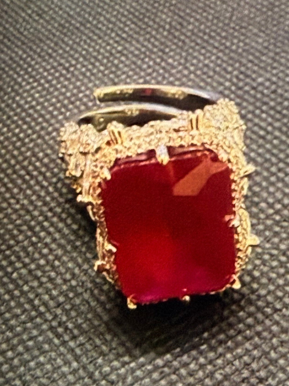 A gold ring with a large square red gemstone, displayed on a dark textured background within a screenshot of a computer screen.