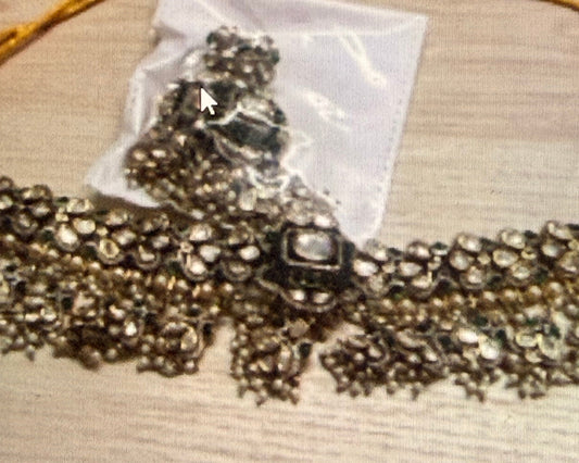 A close-up of an ornate silver-toned beaded necklace or choker laid flat on a light wood surface, with a small clear plastic bag containing matching embellishments or packaging placed on top. The photo appears to be a cropped screenshot showing multiple product photos together.