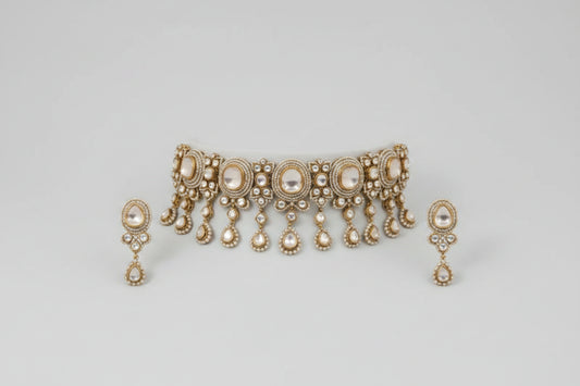 A close-up of a gold-toned traditional Indian jewelry set, including a necklace with large central stones and dangling teardrop motifs, matching earrings, and a bracelet, displayed on a dark fabric background. The photo appears to be a screenshot from a spreadsheet or catalog page, with browser tabs and a green color swatch visible around it.