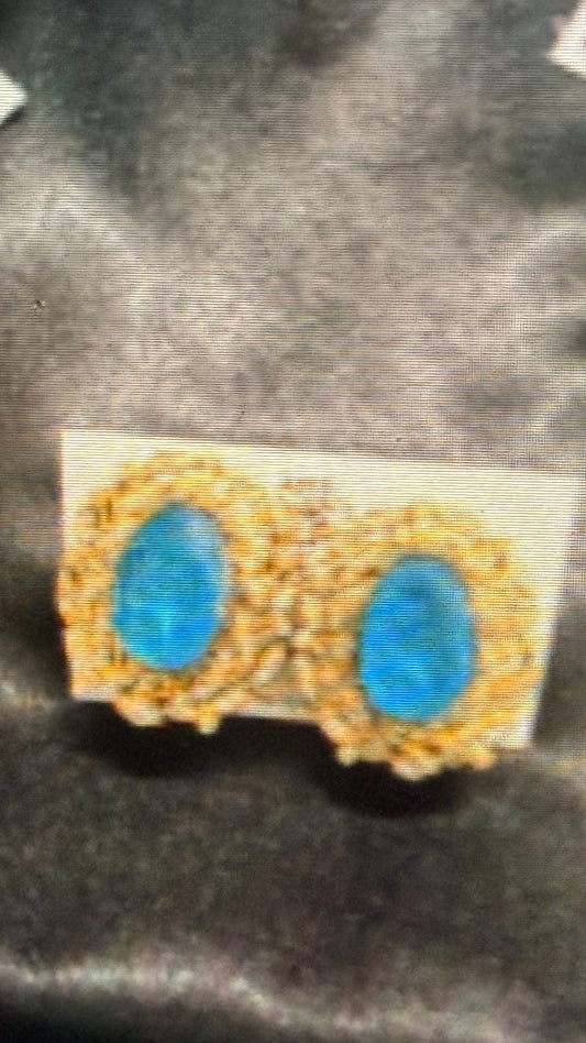 A pair of gold-toned earrings with oval blue stones, displayed on a small white card against a dark satin-like background. The photo appears to be a screenshot of a product listing, with a Windows taskbar visible at the bottom.