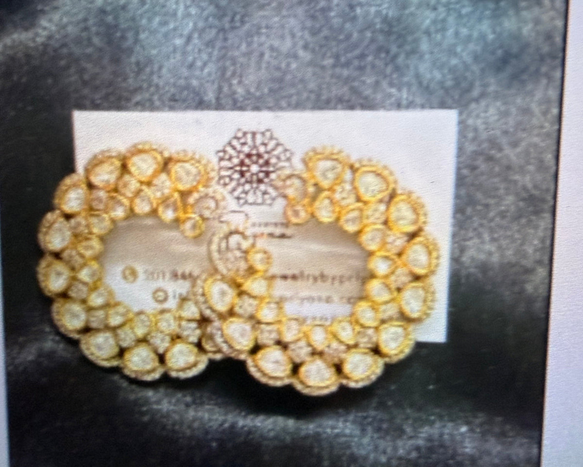 A pair of gold-toned traditional Indian earrings with embedded clear stones, displayed on a white card against a dark textured background.