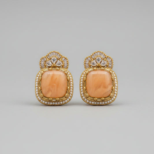 A pair of gold-toned statement earrings with large peach-orange oval cabochons and small clear stones, displayed on a white card. The card is photographed on a dark surface, with a computer monitor partially visible around the edges.