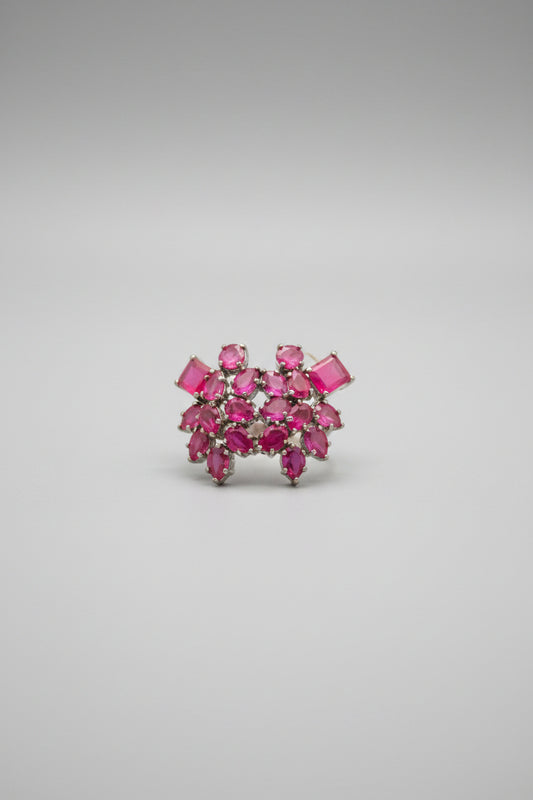 A close-up of a jewelry piece with bright pink stones set in a dark metal, attached to a white branded card, displayed on a dark satin-like fabric.