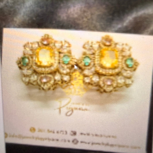 A pair of ornate gold-toned statement earrings with rectangular yellow center stones and small green accent stones, displayed on a white jewelry card labeled 'Priyana'.